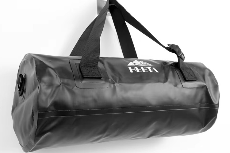 The image features a black waterproof duffel bag with the "HEETA" label on it. It has sturdy black straps and a minimalist design.
