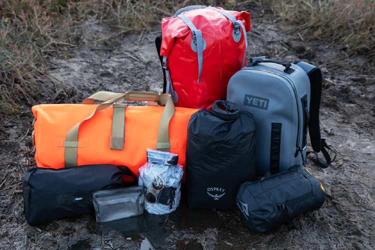  The image shows a variety of outdoor gear including backpacks, dry bags, and other equipment. Brands visible include YETI, Osprey, and others. The items are placed on a muddy ground, suggesting they are suitable for rugged outdoor conditions.