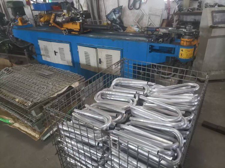 Aluminum surfboard rack tubes stacked in a metal bin beside a tube bending machine in workshop