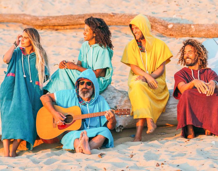 Group of five people wearing colorful surf ponchos on the beach, sitting on driftwood at sunset