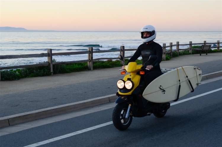 Surfer riding a yellow moped with surfboard mounted on side rack at the beach road