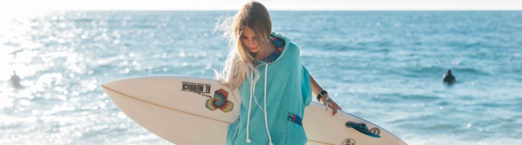 Woman in a turquoise surf poncho holding a surfboard by the ocean