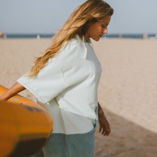 Side profile of woman wearing two-tone surf poncho near paddleboard on sandy beach
