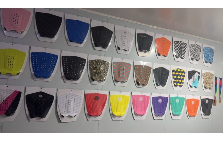 The image shows a collection of surf pads displayed on a wall. The surf pads come in a variety of colors, including pink, black, blue, yellow, green, brown, red, white, and multicolored patterns.
