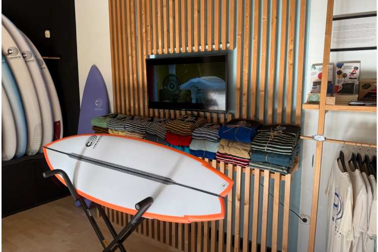 The image depicts a store setting with surfboards on the left and a variety of surfboard covers neatly arranged on a table in the center. There is also a television screen showing surfing footage.