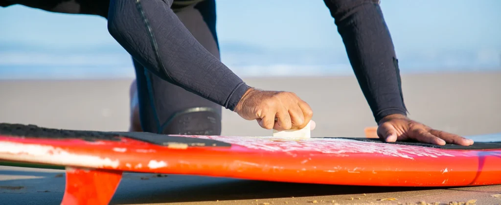 The image shows a person in a wetsuit applying surf wax to a red surfboard on the beach. The person is using their hand to spread the wax evenly across the surface of the board, ensuring better grip for surfing.