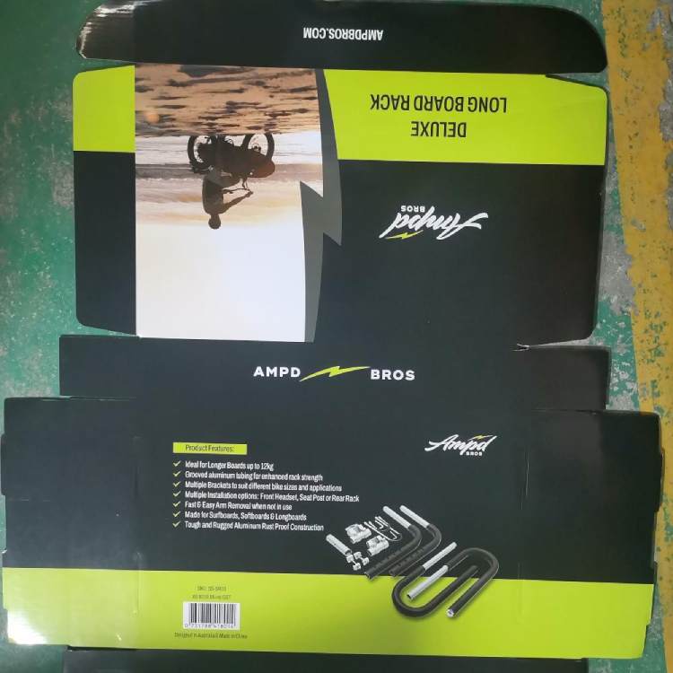 The image shows the packaging of the Ampd Bros custom surfboard bike rack. The packaging is primarily black and green with the brand name "Ampd Bros" prominently displayed. It features product images and a list of product features, such as suitability for longer boards up to 29kg, adjustable aluminum tubing for enhanced rack strength, multiple brackets for different bike sizes, and easy arm removal when not in use. The packaging also highlights that the product is suitable for surfboards, stand - up paddleboards, and longboards, and mentions the tough and rugged aluminum construction.
