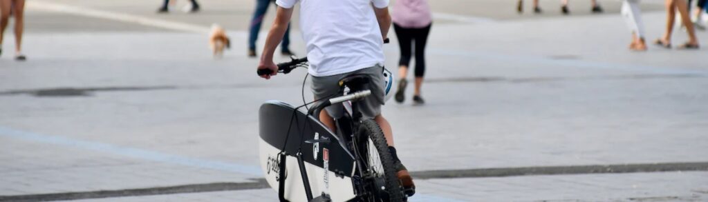 A person is riding a bicycle equipped with a surfboard rack. The rack is attached to the rear of the bike and holds a surfboard securely. The scene takes place in an urban area with other pedestrians and a small dog visible in the background.