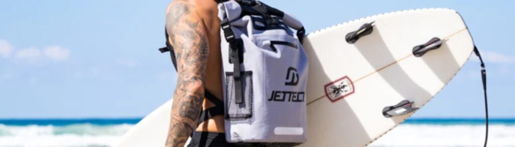 A person is carrying a backpack while standing next to a bicycle that has a surfboard attached to it. The surfboard is blue with a visible logo, and the scene takes place outdoors with trees and a paved path in the background.