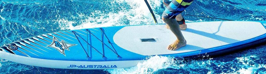 : A person is engaged in stand - up paddleboarding (SUP) on a blue and white JP - Australia paddleboard. In the top left corner, there are two blue pump adapters with black caps, likely used for inflating the paddleboard. The person is holding a paddle and navigating through the water.