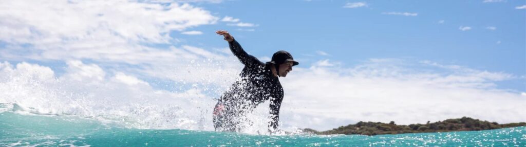 A surfer is captured mid - action, riding a wave with skill. The surfer is wearing a black wetsuit and a cap, with one arm extended for balance. The wave is splashing around them, and the background features a clear blue sky with scattered clouds and a distant shoreline with greenery.
