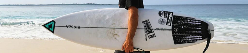 A person is carrying a surfboard on a sandy beach with the ocean and waves in the background. The surfboard has various brand logos and designs on it, including "VZSSIA" and "Futures". The scene captures a typical day at the beach for a surfer.
