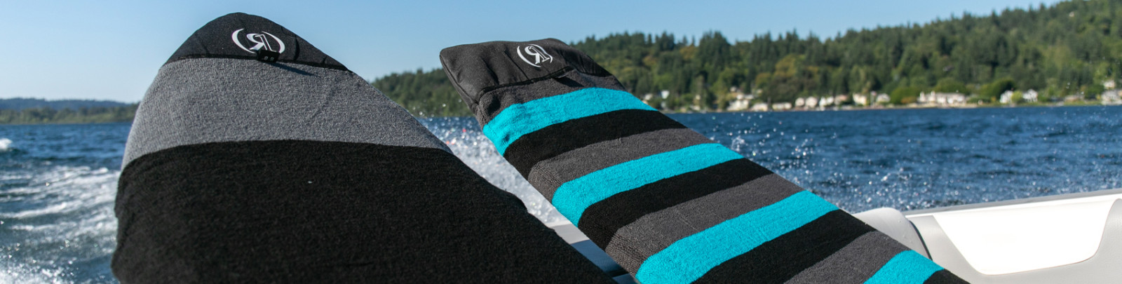 The image shows two surfboard socks with color - blocked patterns. One towel features black and gray sections, while the other has black and teal sections. Both towels have a circular logo on them. The background depicts a body of water with a distant shoreline of trees.